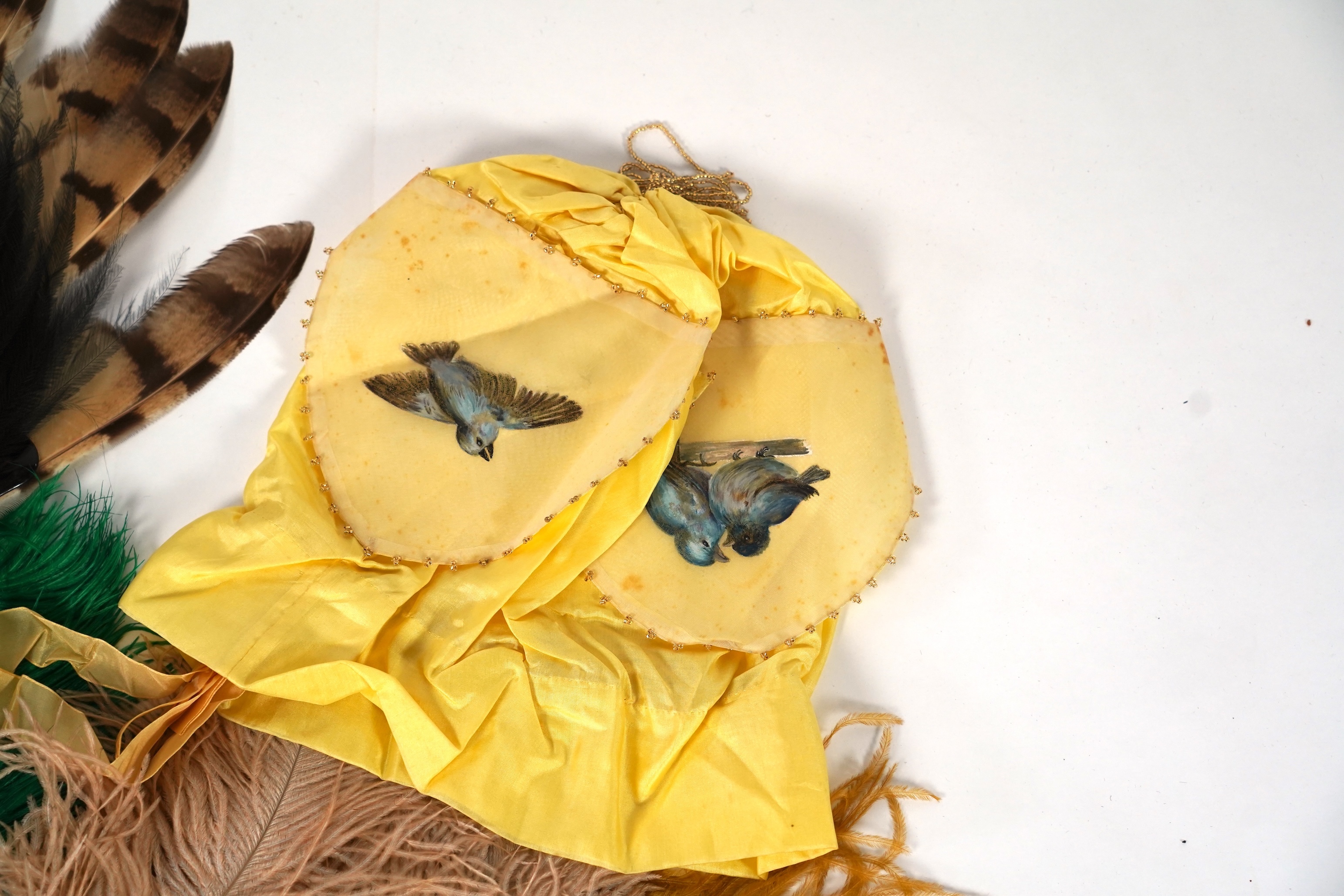 A collection of Edwardian exotic feather hat ornaments, with a collection of coloured ostrich feathers, two similar feather fans, a lace and another fan and silk hand painted mats